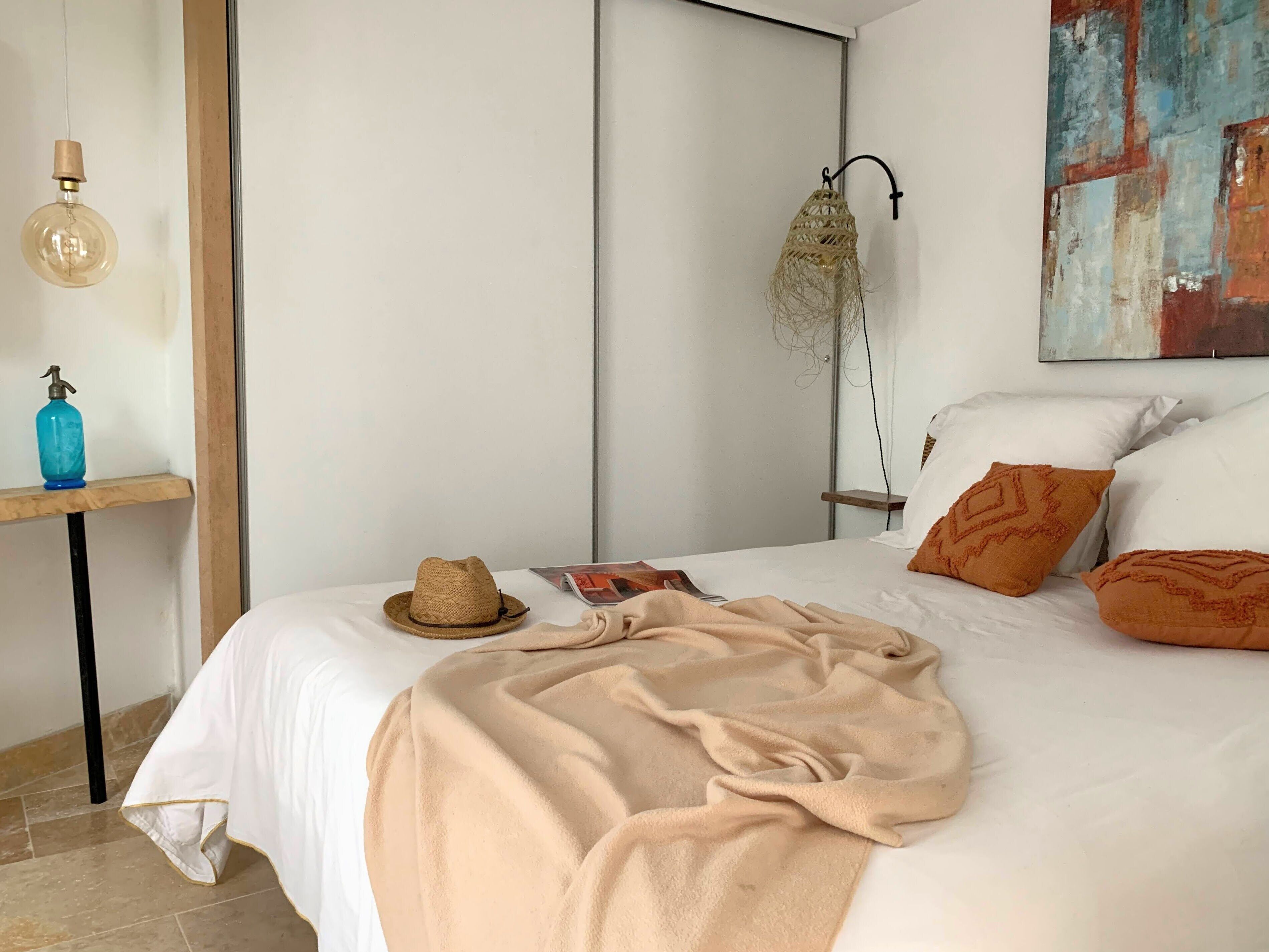 A neatly made bed with a beige blanket, a straw hat, and a magazine on the nightstand.