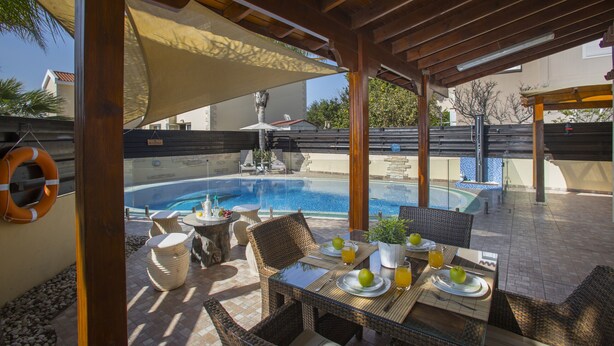 Un área de piscina exterior con un patio cubierto, muebles de mimbre y una mesa lista para comer.