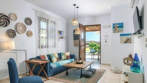 A living room with a sofa, coffee table, and a TV. There are decorative items on the walls and a view of the outdoors. - Lindos Topaz - Two Bedroom Villa, Sleeps 4 (Lindos)