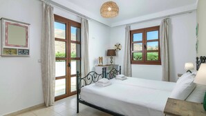 A bedroom with a bed, curtains, a hanging light fixture, a bedside table, and a window with a view of greenery. - Lindos Topaz - Two Bedroom Villa, Sleeps 4 (Lindos)
