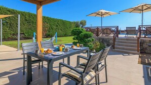 An outdoor patio with a table set for breakfast, surrounded by chairs and umbrellas, overlooking a landscaped area with a pool and stairs leading to a wooden deck. - Lindos Topaz - Two Bedroom Villa, Sleeps 4 (Lindos)