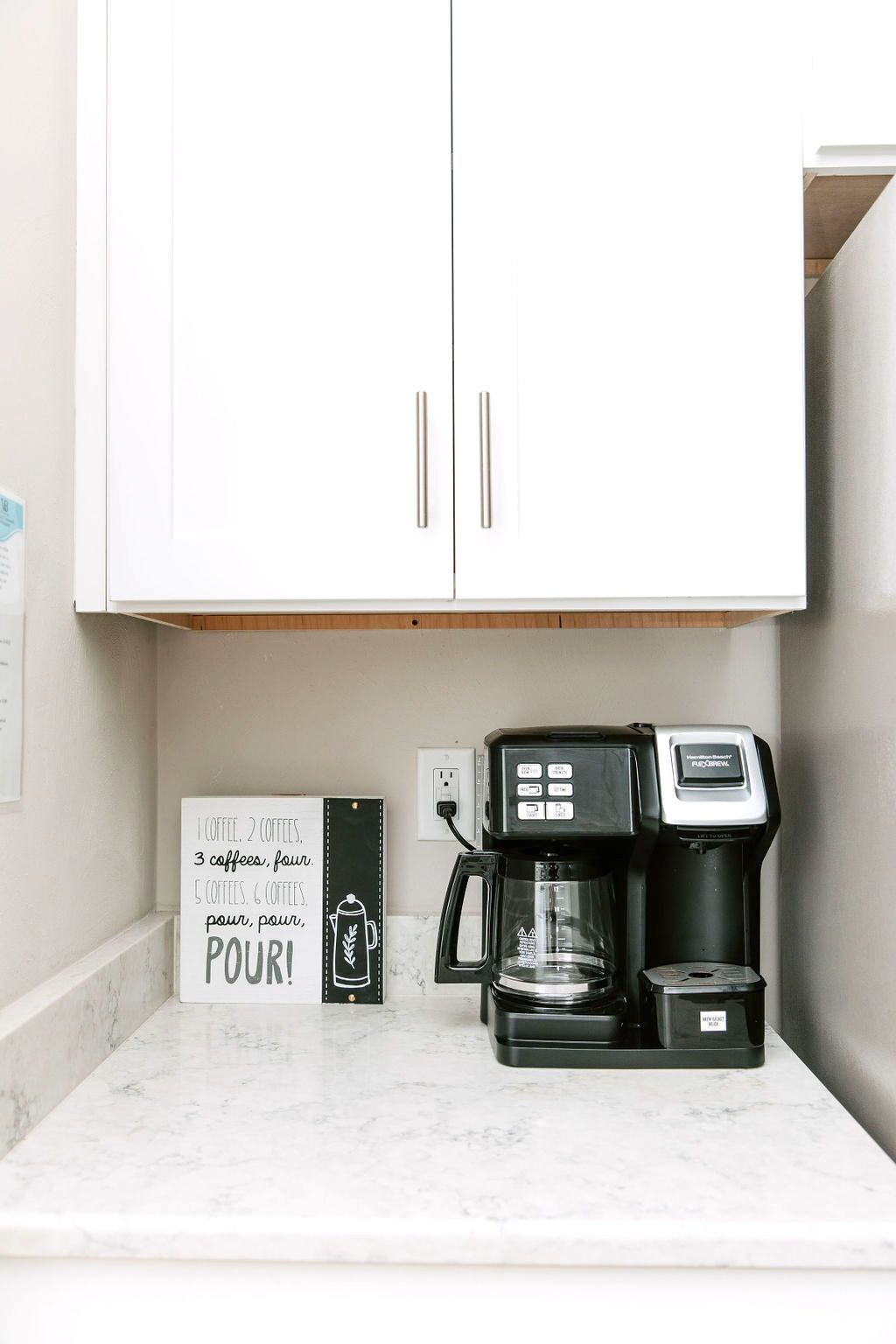 Un coin cuisine équipé d’une machine à café, d’une pancarte avec des informations sur le café, et d’un meuble blanc placé au-dessus.