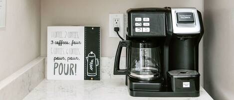 Un coin cuisine équipé d’une machine à café, d’une pancarte avec des informations sur le café, et d’un meuble blanc placé au-dessus.