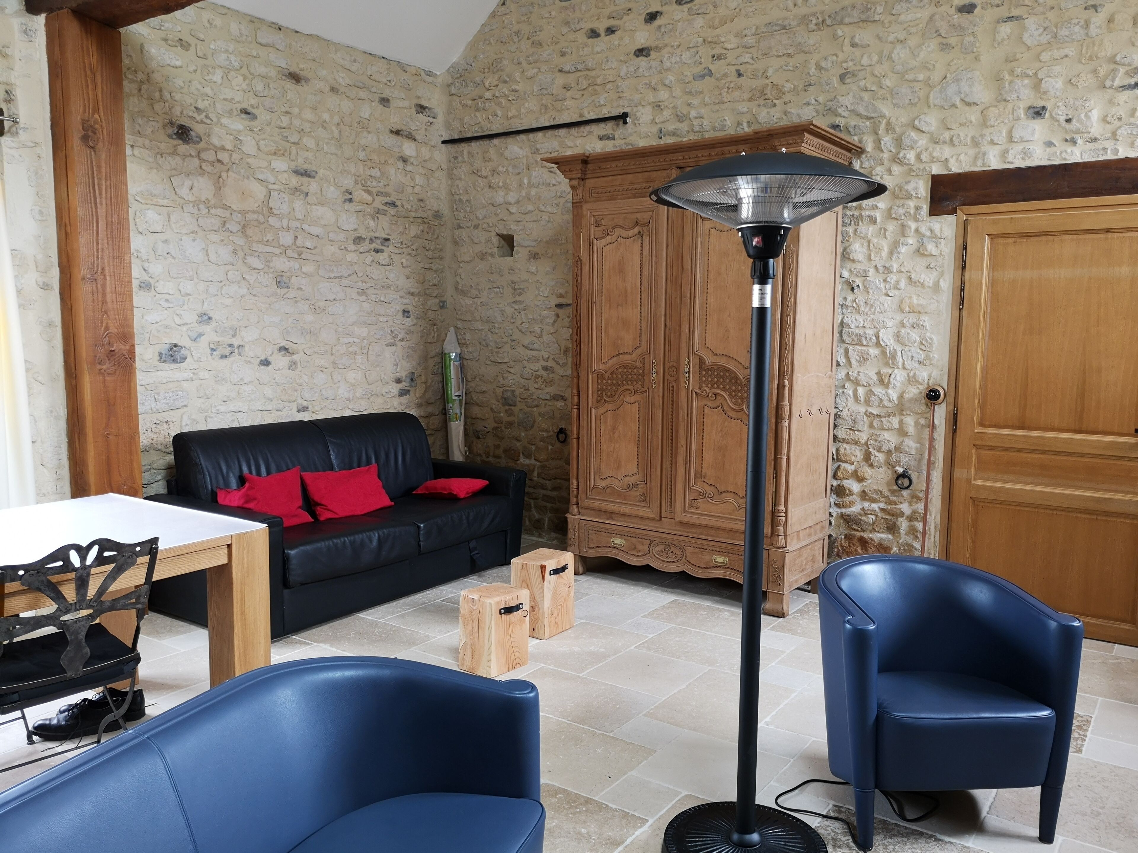 A room with a stone wall, a black leather sofa with red cushions, a wooden cabinet, a tall floor lamp, and two blue chairs.