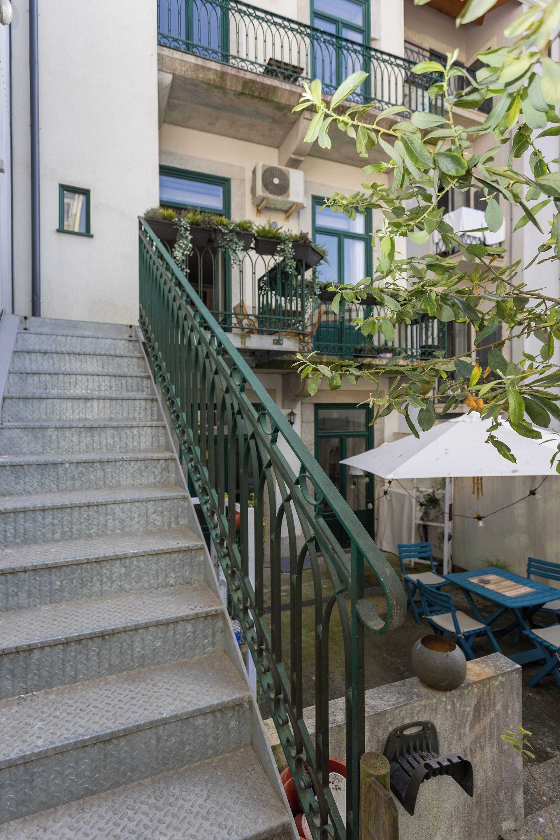A staircase with a metal railing leading to a building with balconies. There are outdoor tables and chairs, and a potted plant is visible.