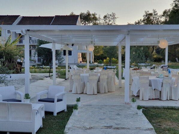 An outdoor event space with white tables and chairs, a covered walkway, and potted plants.