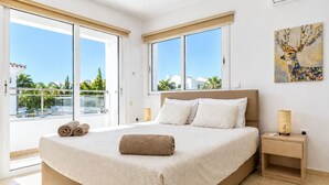 A hotel room with a large bed, two windows, a painting on the wall, and a balcony with a view of palm trees and buildings. - Narcissos Villa 3 - Three Bedroom Villa, Sleeps 8 (Protaras)