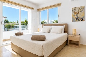 Hotelkamer met een groot bed, twee ramen, een schilderij aan de muur en een balkon met uitzicht op palmbomen en gebouwen.