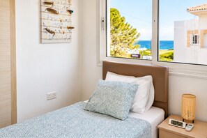 A bedroom with a bed, a wall-mounted artwork, a bedside table with a lamp, and a view of the sea through the window.