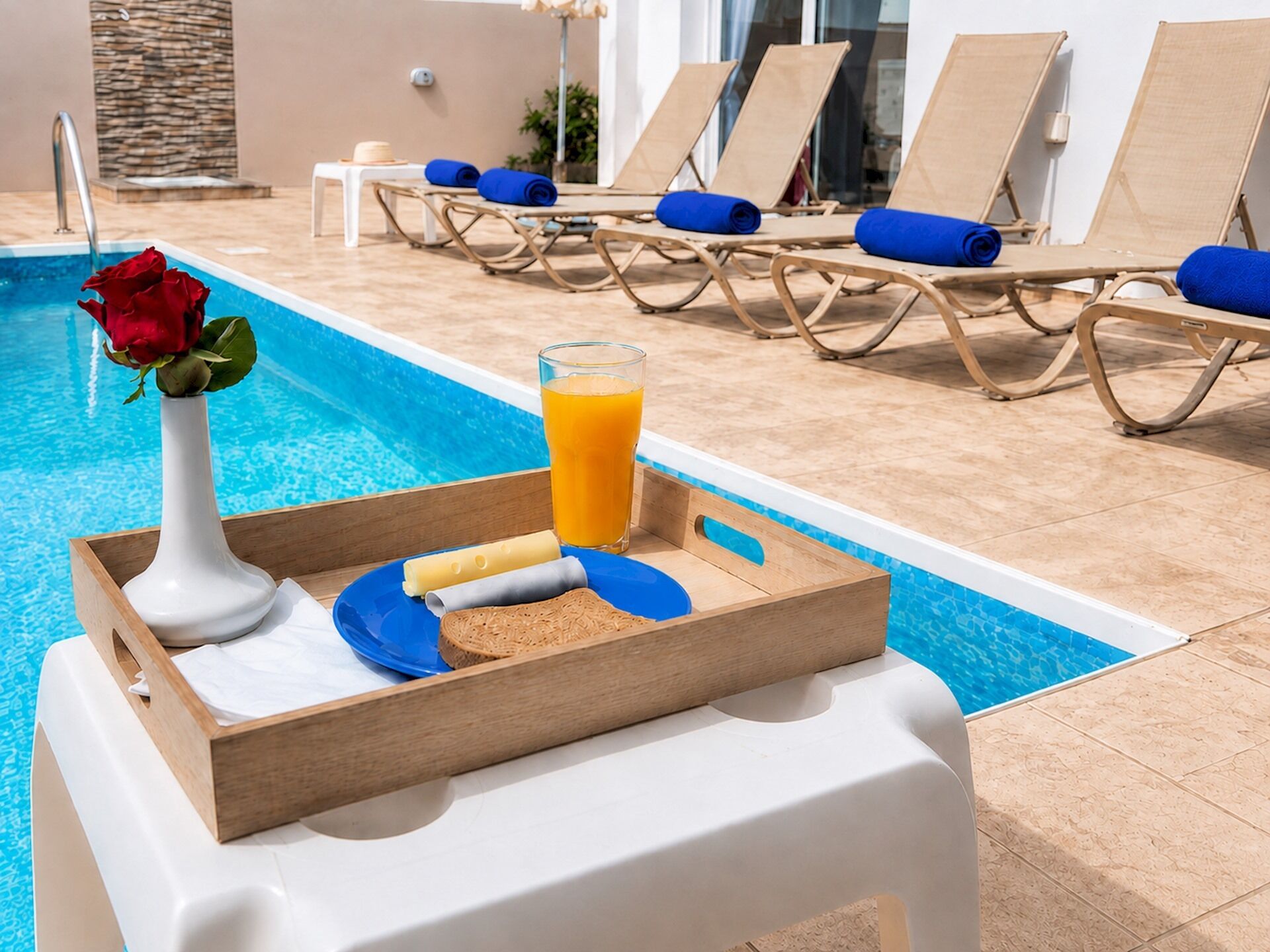 A poolside setting with lounge chairs, a tray with food and a drink, and a rose in a vase.