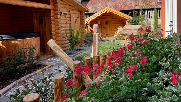 Eine Blockhütte mit einem Steinpfad, der zu einer Holzdiele führt, umgeben von einem Garten mit roten Blüten.