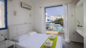 Ein Hotelzimmer mit Bett, einem kleinen Tisch mit Pflanze, einem Balkon mit Pool und Blick auf Gebäude.