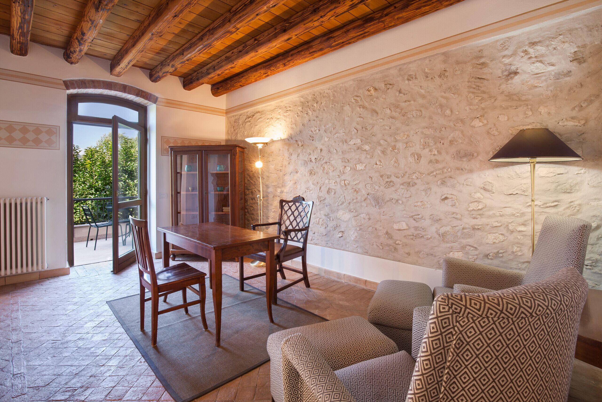 A cozy room with a stone wall, wooden ceiling beams, a dining table with chairs, a patterned sofa, and a floor lamp.