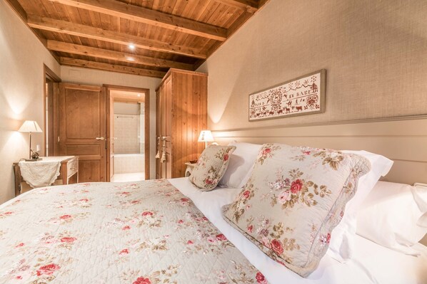 A bedroom with a wooden ceiling, a bed with floral bedding, a nightstand with a lamp, and a door leading to another room. - Val de Ruda Luxe 11 by FeelFree Rentals (Baqueira)