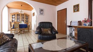 Una sala de estar con techo de madera, un sofá de cuero negro, una mesa de centro de vidrio, un comedor con mantel azul y un mueble con puertas de cristal.