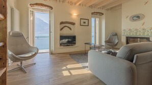 A living room with a sofa, two chairs, a TV, and a fireplace. There are two doors leading to balconies with views of mountains. - Elizabeth House Varenna (Varenna)