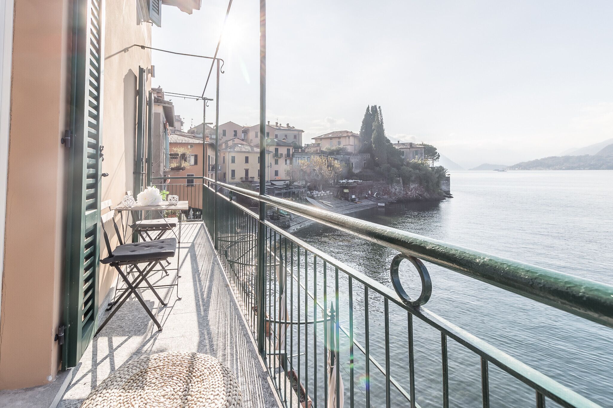 A balcony with a view of a calm lake, buildings in the background, and a railing.
