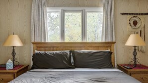 A bedroom with a wooden headboard, two bedside tables with lamps, a window with curtains, and a wall tapestry.