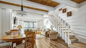 A spacious living room with a wooden dining table, comfortable seating, a staircase, and a large window providing natural light.
