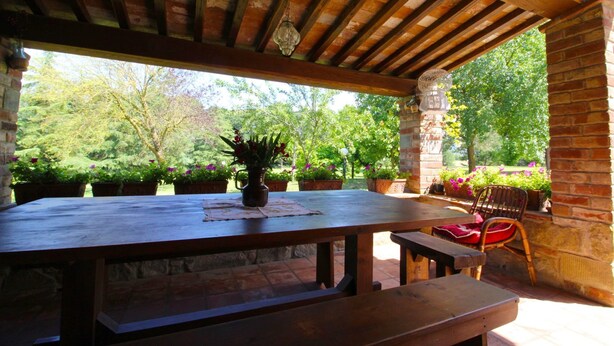 Una terrazza esterna con tavolo e panche di legno, circondata da piante in vaso e un muro in mattoni.