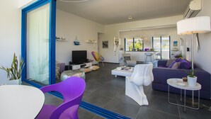 A modern living room with a sofa, chairs, a TV, and a dining area. - Protaras Aster Seaside Suite (Protaras)