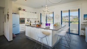 A modern kitchen with a central island, bar stools, a chandelier, and a view through large windows. - Protaras Aster Seaside Suite (Protaras)