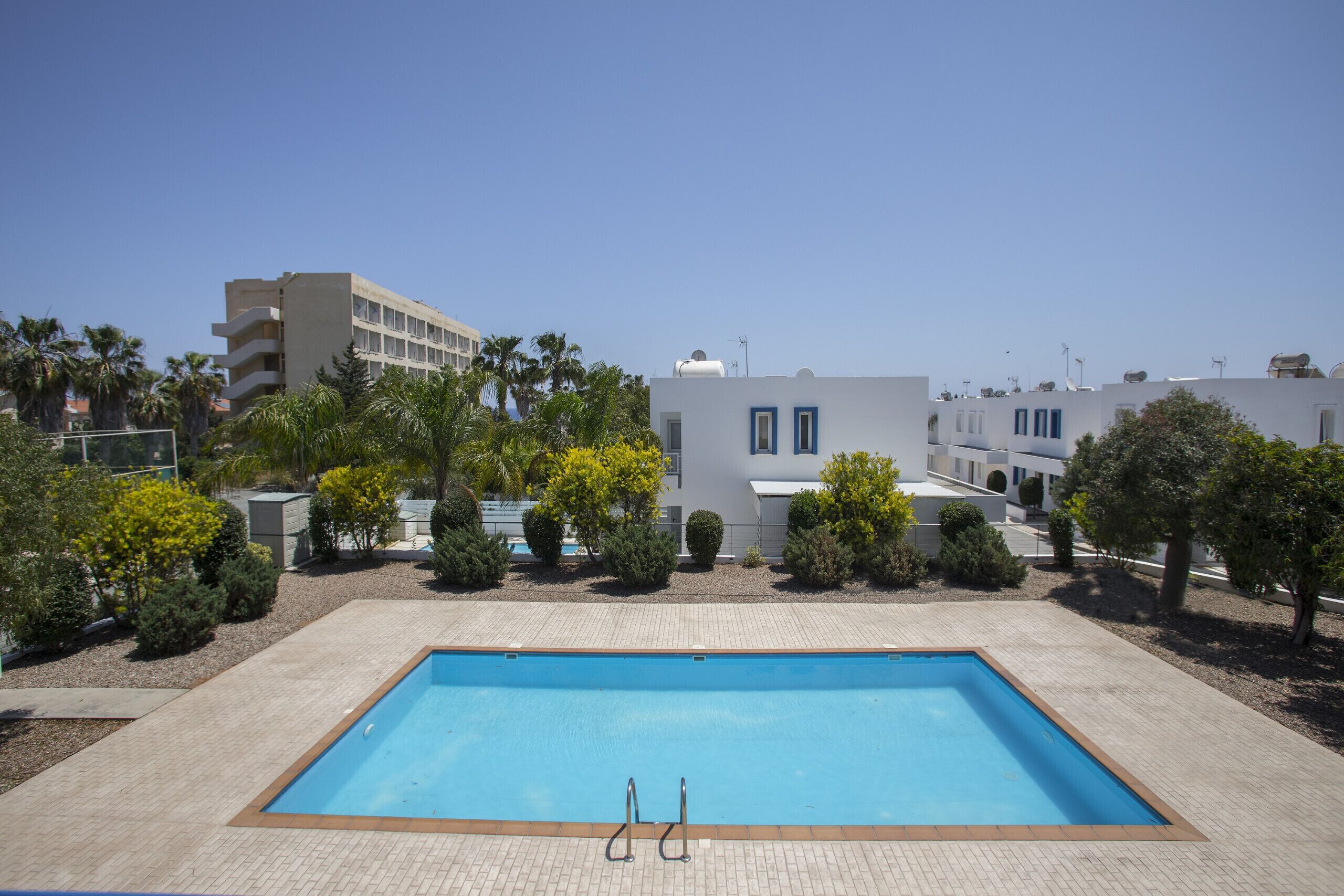 Una piscina rectangular amb dues escales, envoltada per una zona pavimentada amb arbres petits i arbustos, i edificis al fons.