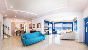 Een ruime woonkamer met een blauwe bank, een witte bank en een eettafel. Achtergrond is een keuken en er is uitzicht op de zee door grote ramen.