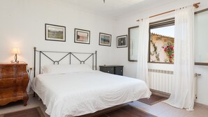 Un dormitorio con cama, cómoda de madera y mesita de noche. Hay tres cuadros enmarcados en la pared, una ventana con cortinas y un radiador.