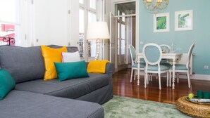 A living room with a grey sofa, teal and yellow cushions, a green rug, and a dining area with white chairs and a table.