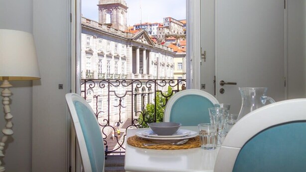 A dining area with a table set for two, a view of a clock tower, and a balcony with a wrought iron railing.