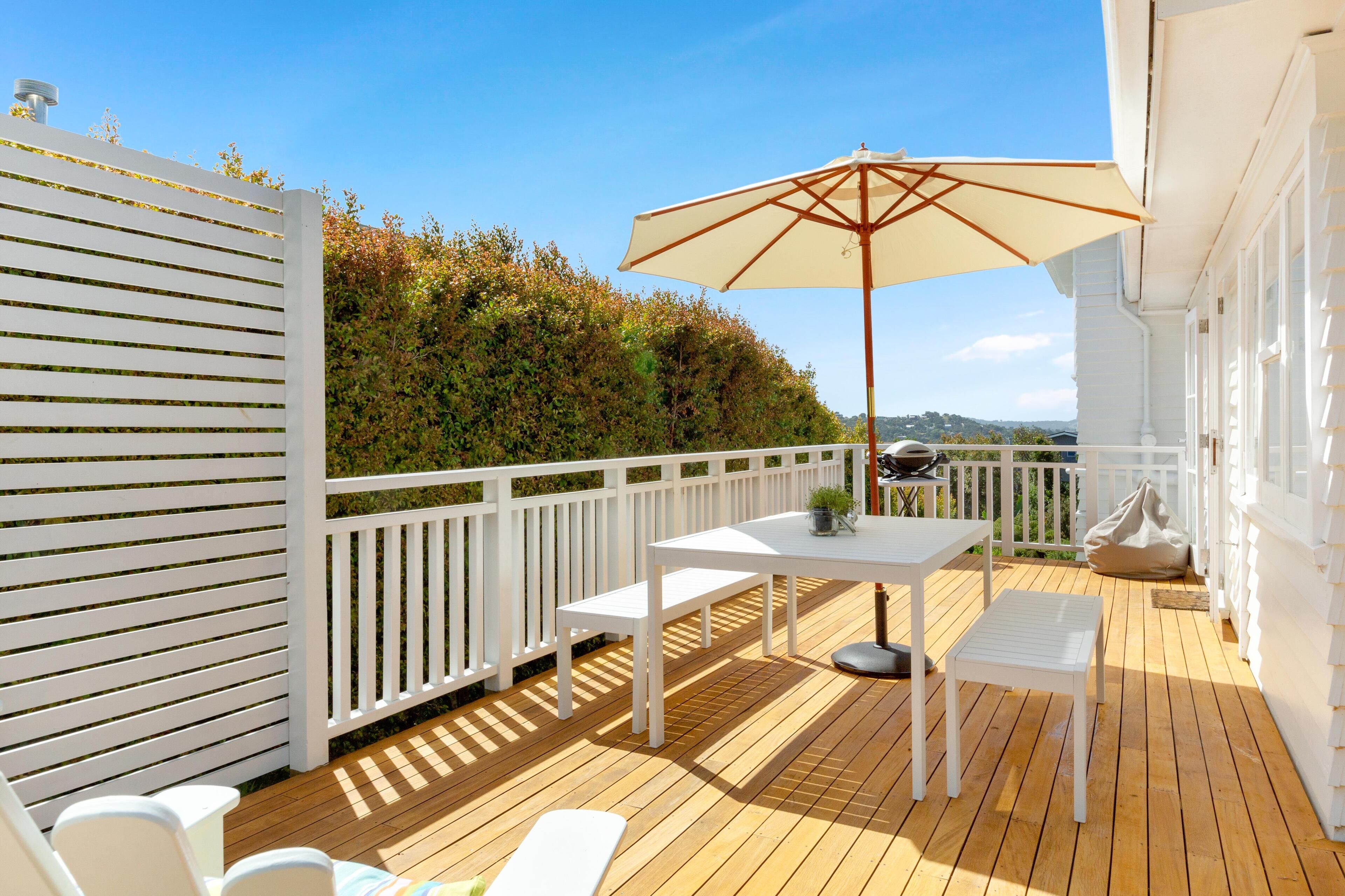 Una terraza de madera con una mesa y banco blancos, un paraguas y una valla blanca.