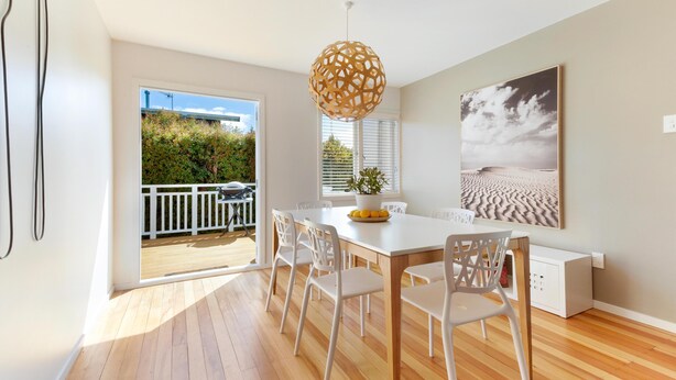 Un comedor moderno con una mesa y sillas blancas, suelo de madera y un ventanal con vistas a zonas verdes y una valla blanca.