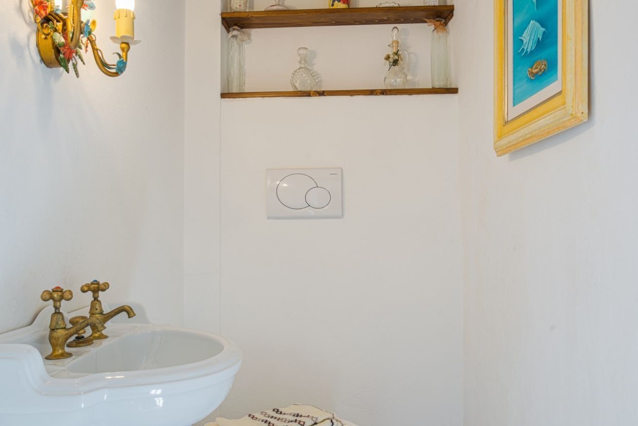 A bathroom with a white sink, brass faucets, a wall-mounted light fixture, and a shelf with glass bottles.