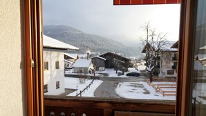 A snowy village scene through a window with wooden shutters, featuring traditional buildings with snow-covered roofs and a clear view of the surrounding hills. - 2-room vacation apartment, approx. 45sqm with sunny south-facing balcony (Reit im Winkl)
