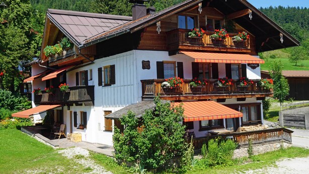 A two-story wooden chalet with balconies, a covered patio, and a mountain backdrop. - 2-room vacation apartment, approx. 45sqm with sunny south-facing balcony (Reit im Winkl)