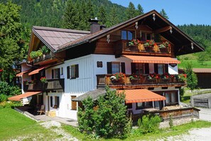 A two-story wooden chalet with balconies, a covered patio, and a mountain backdrop. - 2-room vacation apartment, approx. 45sqm with sunny south-facing balcony (Reit im Winkl)