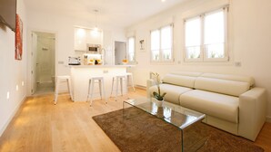 Una sala de estar moderna con una mesa de centro de vidrio, un sofá blanco y una zona de cocina con microondas y taburetes.
