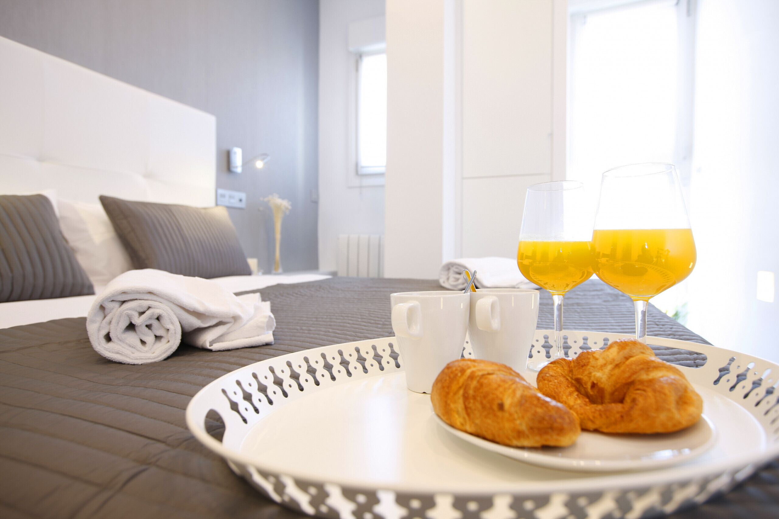 Cama bem arrumada com almofadas, uma toalha dobrada, um prato com croissants e um copo de suco de laranja, e duas canecas brancas sobre uma mesa em quarto de hotel.
