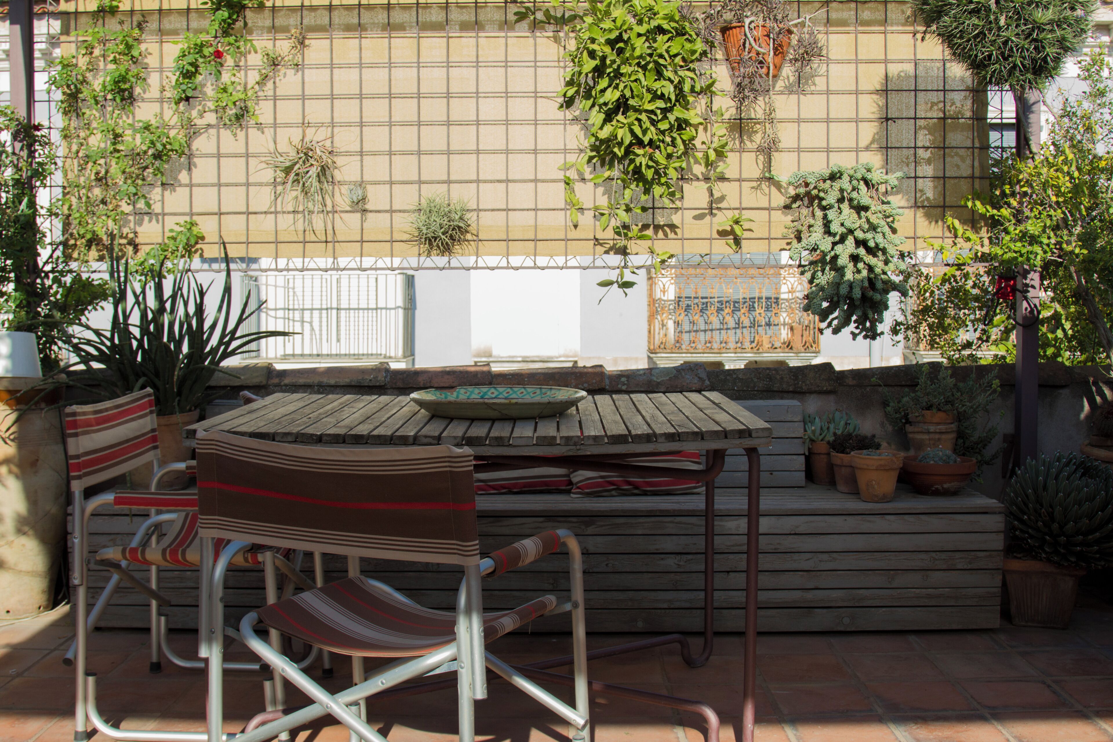 A wooden table and chairs set outdoors with potted plants and a metal fence in the background.