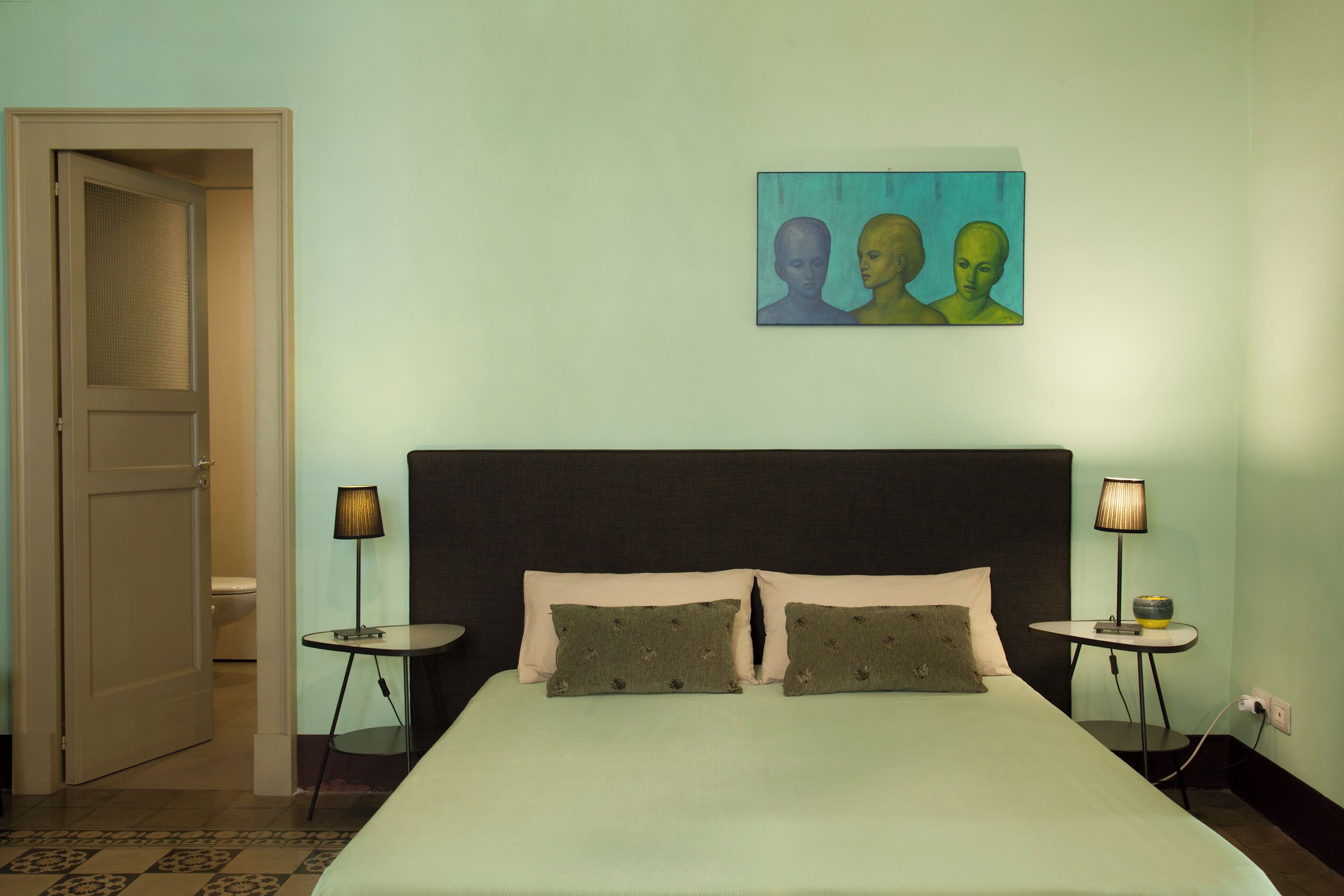 A hotel room with a bed, two bedside tables, a painting, and a door leading to a bathroom.