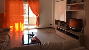A living room with a sofa, a glass coffee table, a TV on a stand, and a sliding glass door with orange curtains. - Paolo Suite in via Parioli in Gela (Gela)