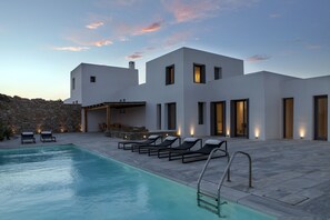 A modern villa with a swimming pool, outdoor seating, and a stone wall backdrop. - NEW LIBERATION VILLA 1 MYKONOS (Mykonos Egeo)