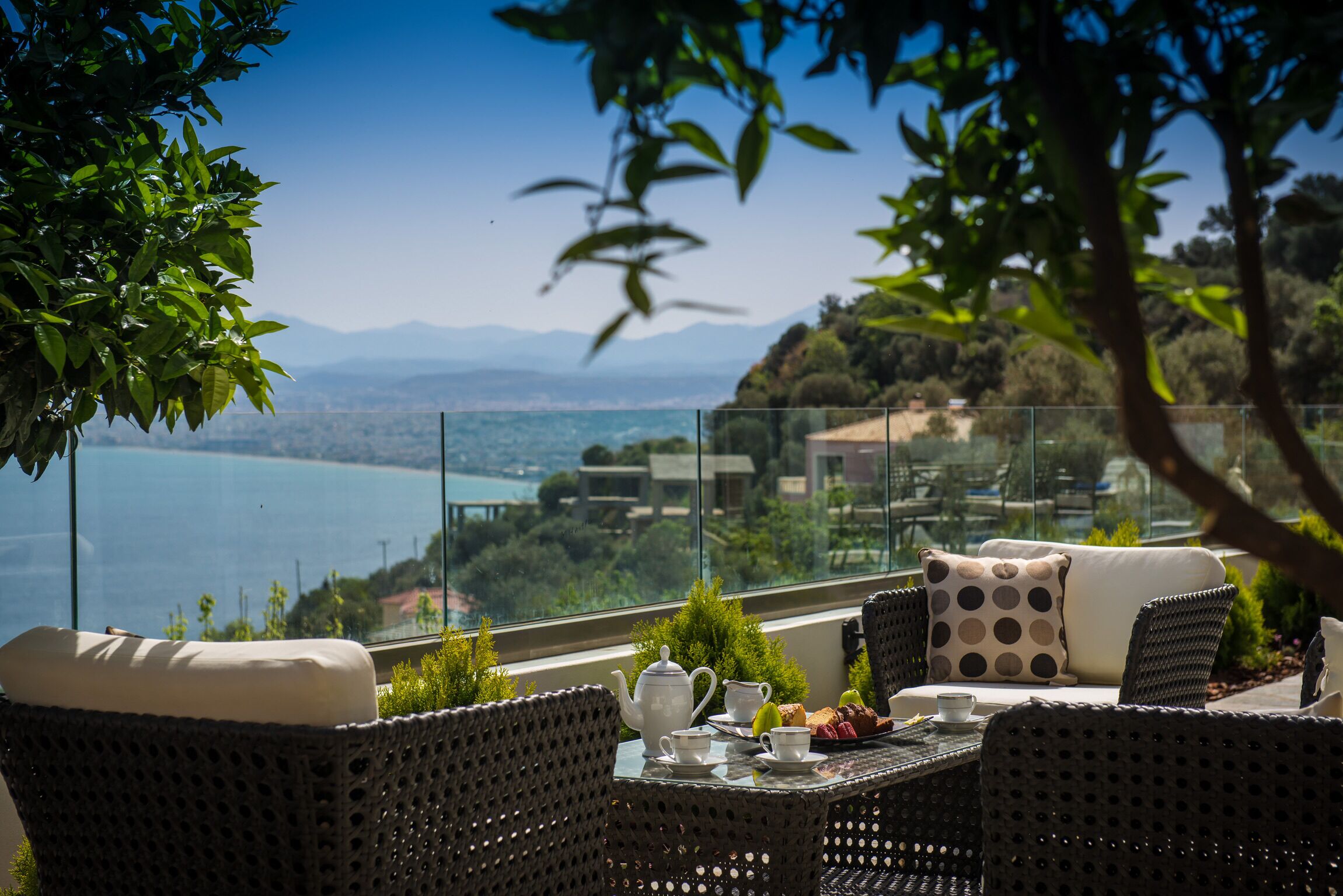 A balcony with wicker furniture, a table with tea set, and a scenic view of the sea and mountains.