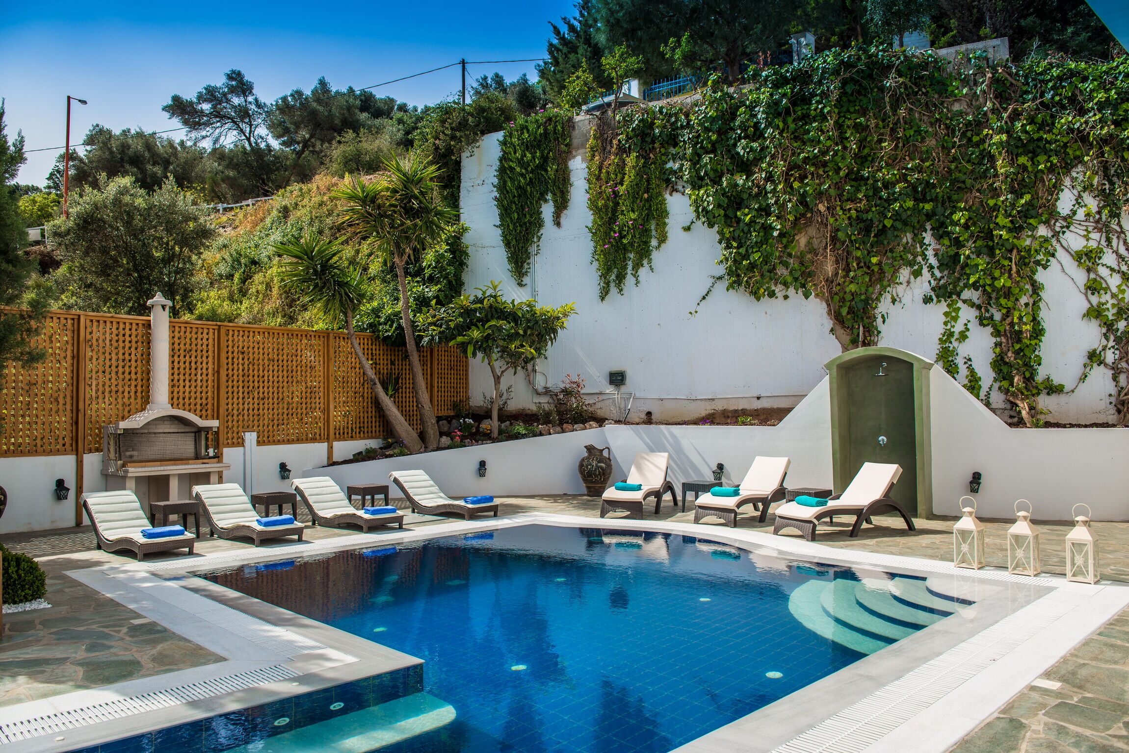 A swimming pool with lounge chairs and a barbecue area, surrounded by greenery and a white wall.