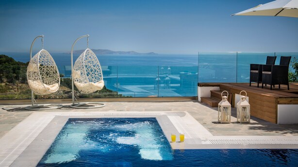 Ein Poolbereich mit zwei weißen Hängestühlen, einer Holzterrasse und Blick aufs Meer.