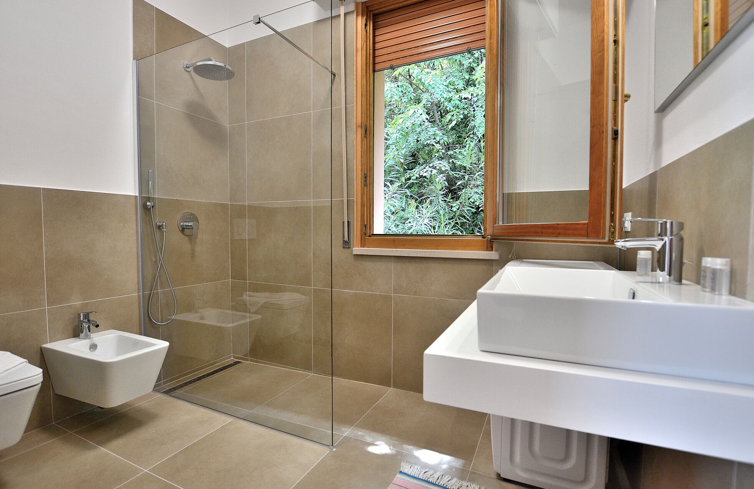 A modern bathroom with a glass-enclosed shower, a white sink, and a window with a view of greenery.