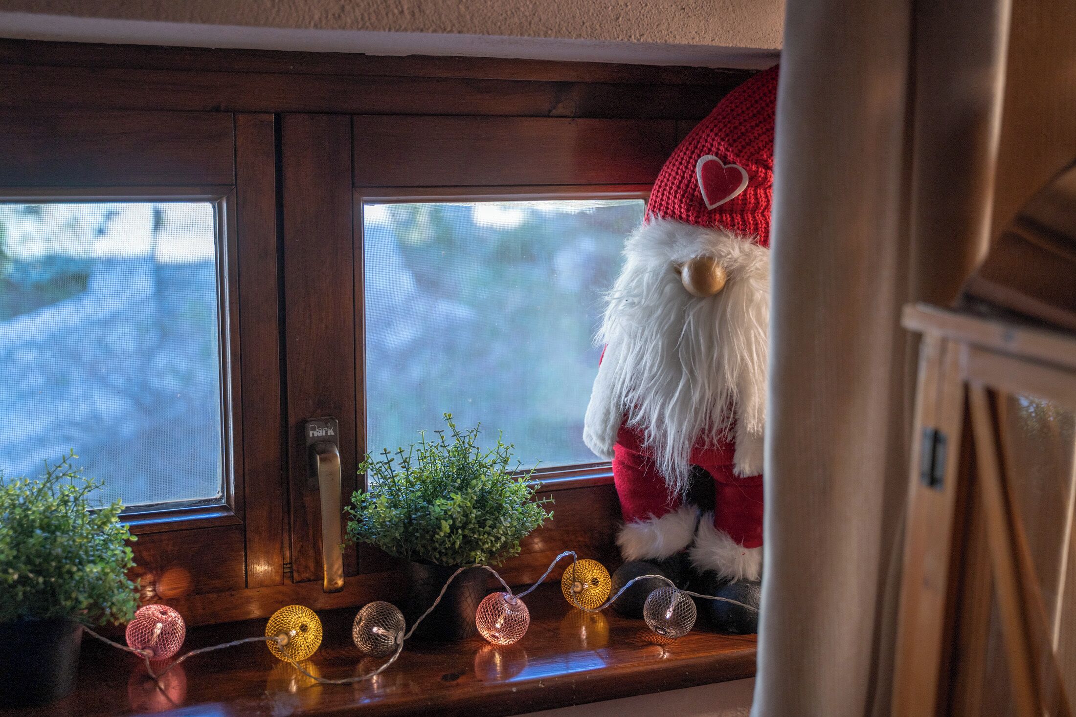 Un rebord de fenêtre orné d’une figurine de Père Noël en peluche, d’une plante en pot et de guirlandes lumineuses décoratives.
