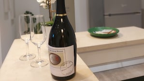 Une bouteille de vin posée sur un comptoir, avec deux verres, une assiette verte et un vase contenant des fleurs blanches.