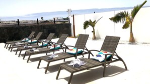 Eine Reihe von Korbsesseln mit Kissen, auf einer Terrasse mit weißer Wand und Blick auf das Meer.